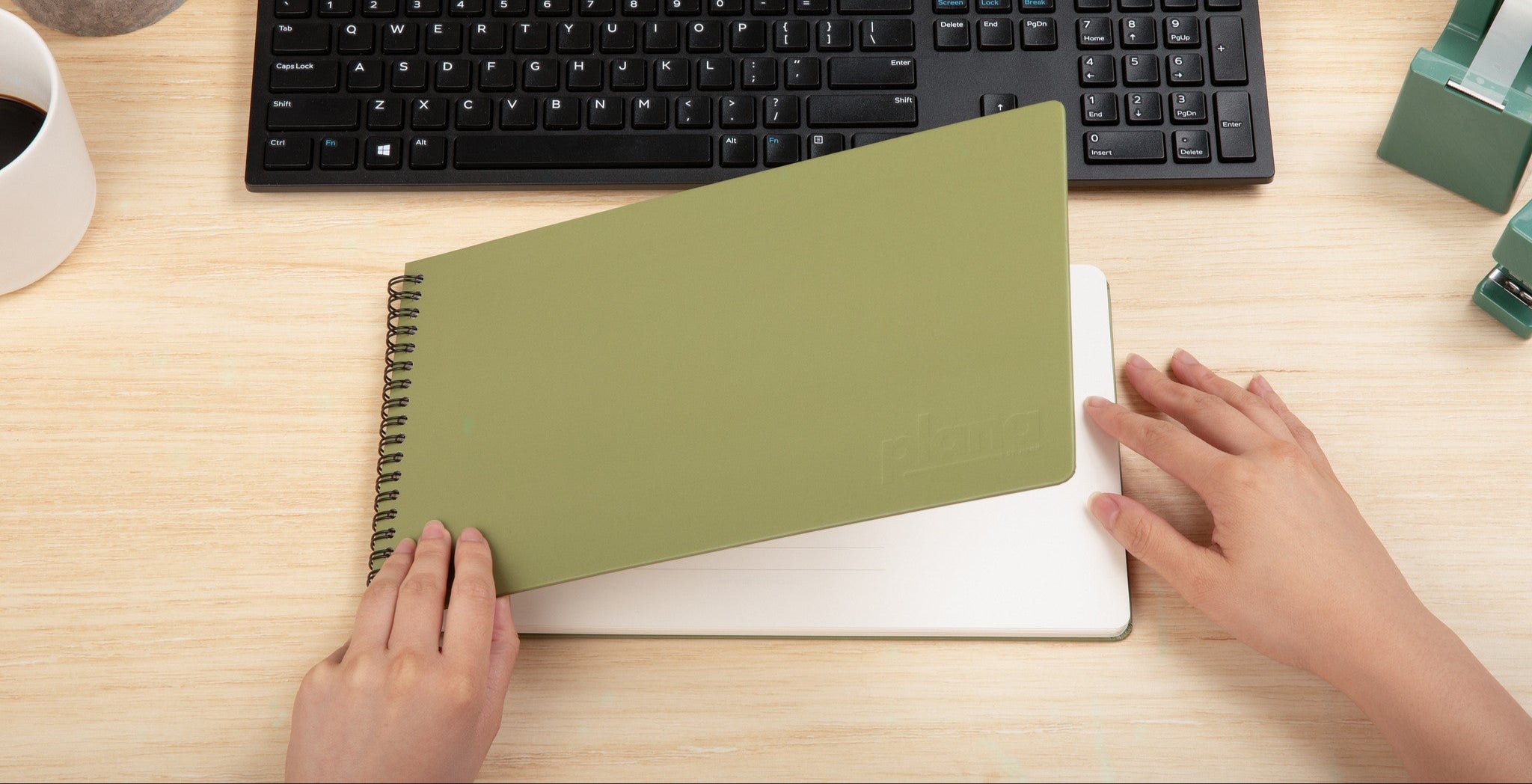 Person using a Plana Classic Cedar notebook on a desk with a keyboard and office supplies.